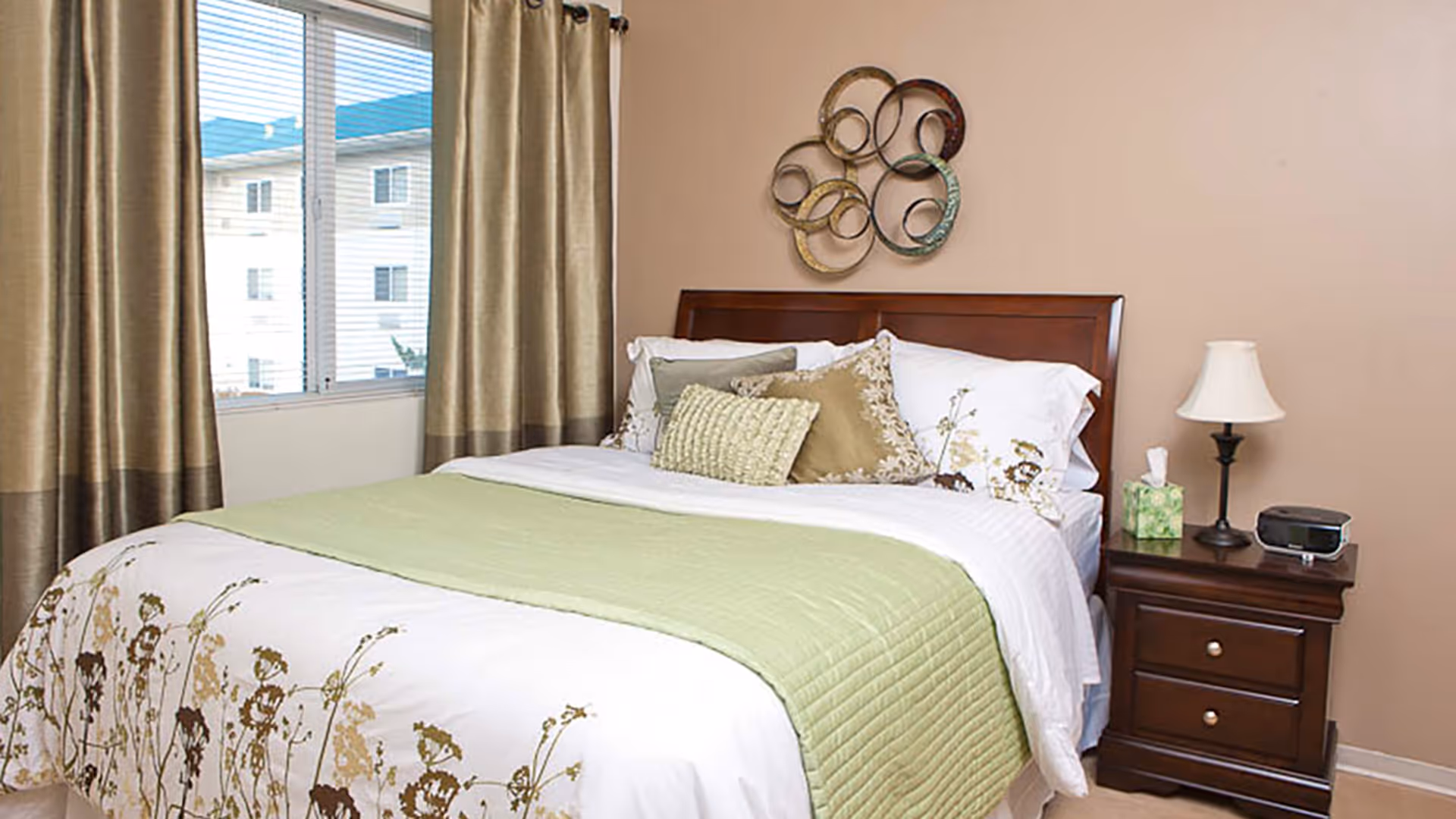 A neatly made bed with white and floral bedding and a light green quilted blanket in a bedroom. There are several decorative pillows on the bed. Next to the bed is a wooden nightstand with a lamp, a tissue box, and a small electronic device. The wall behind the bed is beige with a circular metal wall decoration. A window with beige curtains is visible on the left side.
