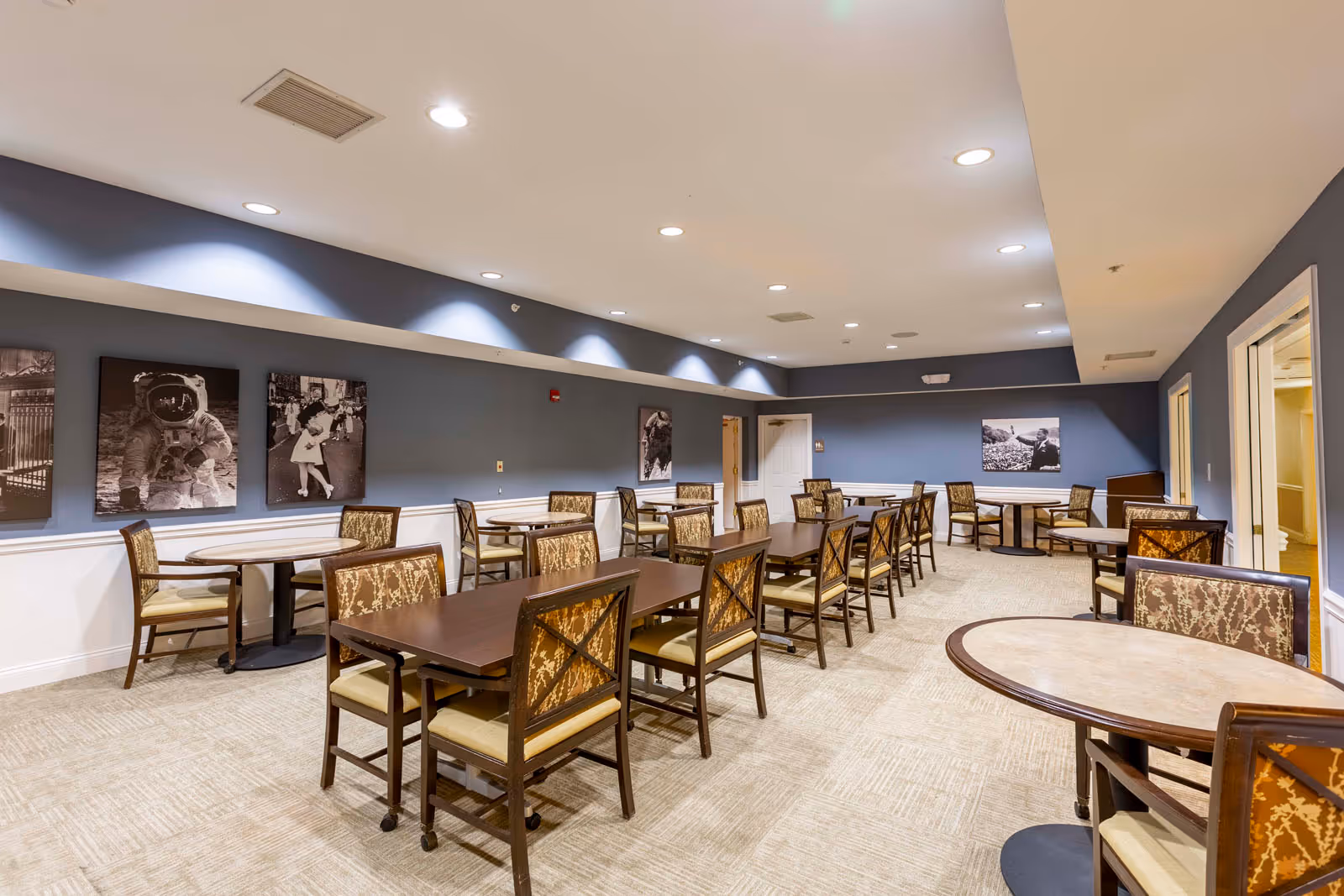 A spacious dining room with multiple tables and chairs arranged neatly. The walls are painted blue with white wainscoting, and black and white photographs are hung on the walls. The ceiling has recessed lighting, and the carpet is beige.