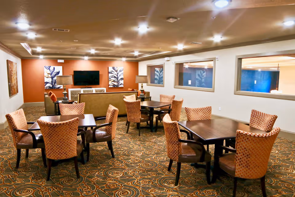 A well-lit common area with multiple dark wood tables surrounded by patterned upholstered chairs. In the background, there is a seating area with a sofa facing a wall-mounted flat-screen TV, flanked by two table lamps and decorative artwork on the walls. The room has a patterned carpet and several windows on one side.