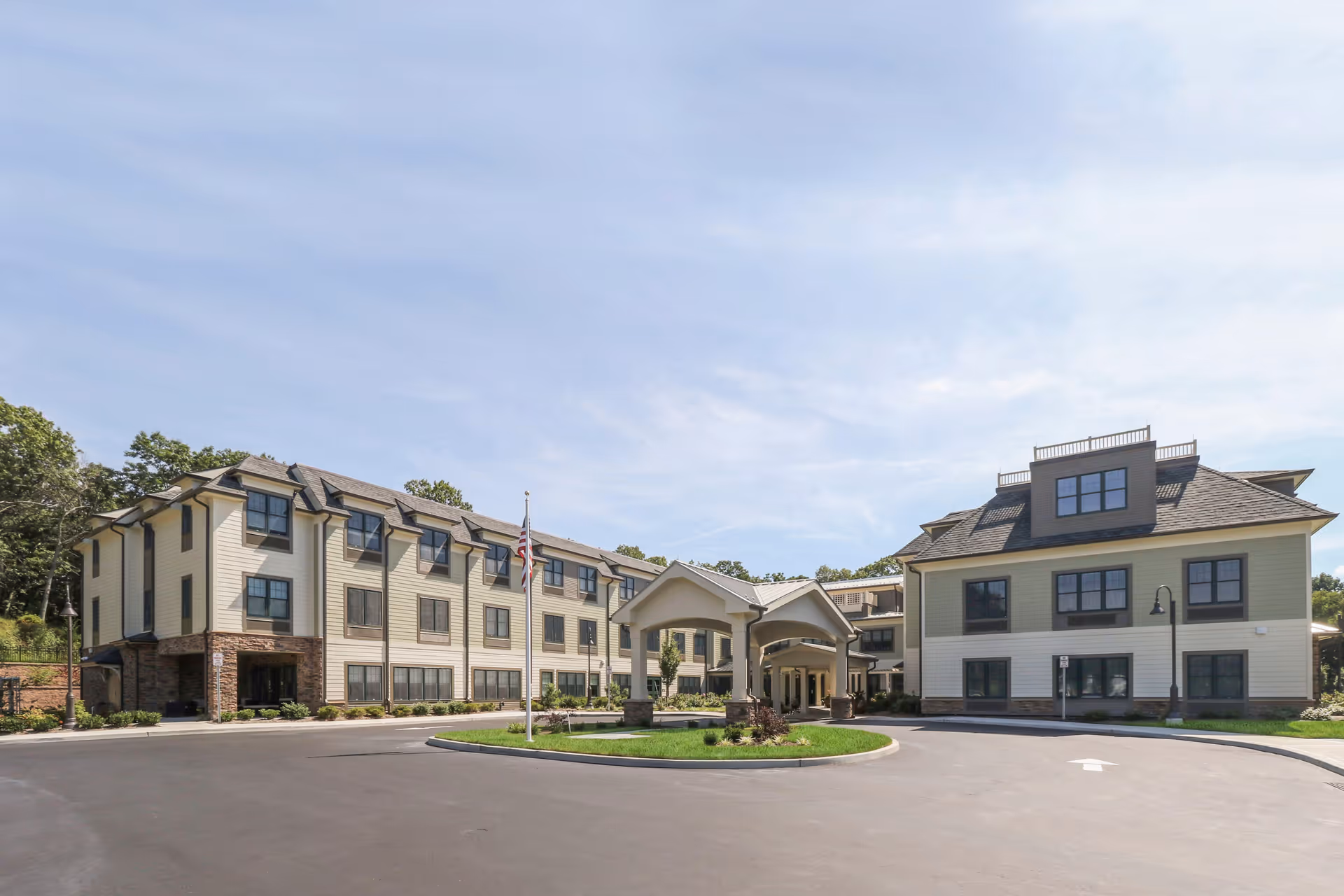 Exterior view of Whisper Woods of Smithtown - Assisted Living & Memory Care facility showing a large, multi-story building with beige siding, multiple windows, a covered entrance, and a circular driveway with a flagpole in front under a partly cloudy sky.