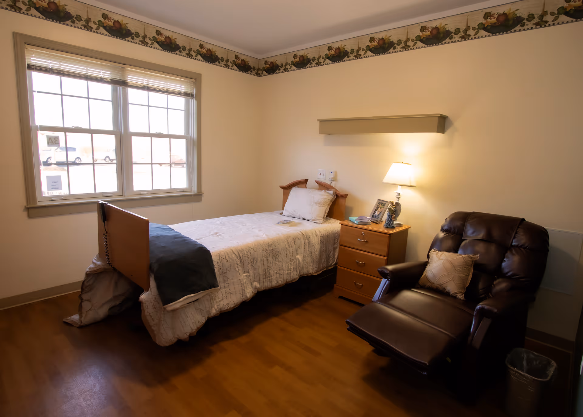 A small, cozy bedroom in a nursing facility with a single bed covered in a white patterned bedspread and a dark blanket at the foot. Next to the bed is a wooden nightstand with a lamp, framed photos, and a figurine. A comfortable dark brown recliner chair with a decorative pillow is positioned nearby. The room has a large window with blinds, light-colored walls with a fruit-themed border near the ceiling, and wood flooring.
