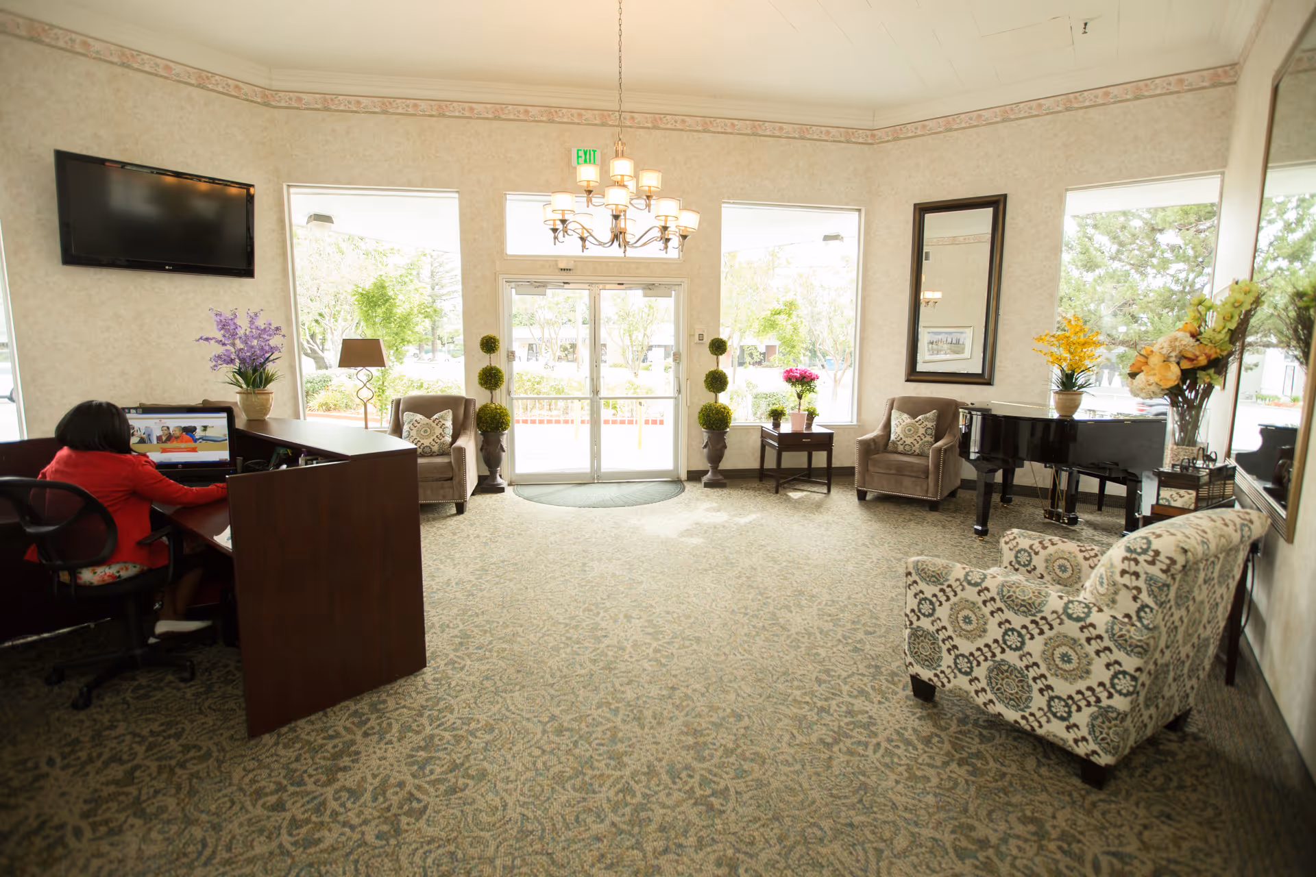 A bright and spacious reception area with a patterned carpet, a chandelier, and large windows letting in natural light. There is a reception desk with a person working on a computer, several armchairs with decorative pillows, a small table with flowers, a grand piano, and various plants and floral arrangements. Double glass doors lead outside.