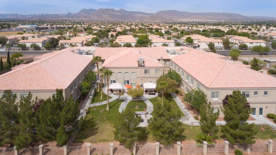Aerial view of Desert View Senior Living facility showing a large, U-shaped building with a tiled roof surrounding a green courtyard with trees, pathways, and seating areas. The facility is located in a suburban area with mountains visible in the background.