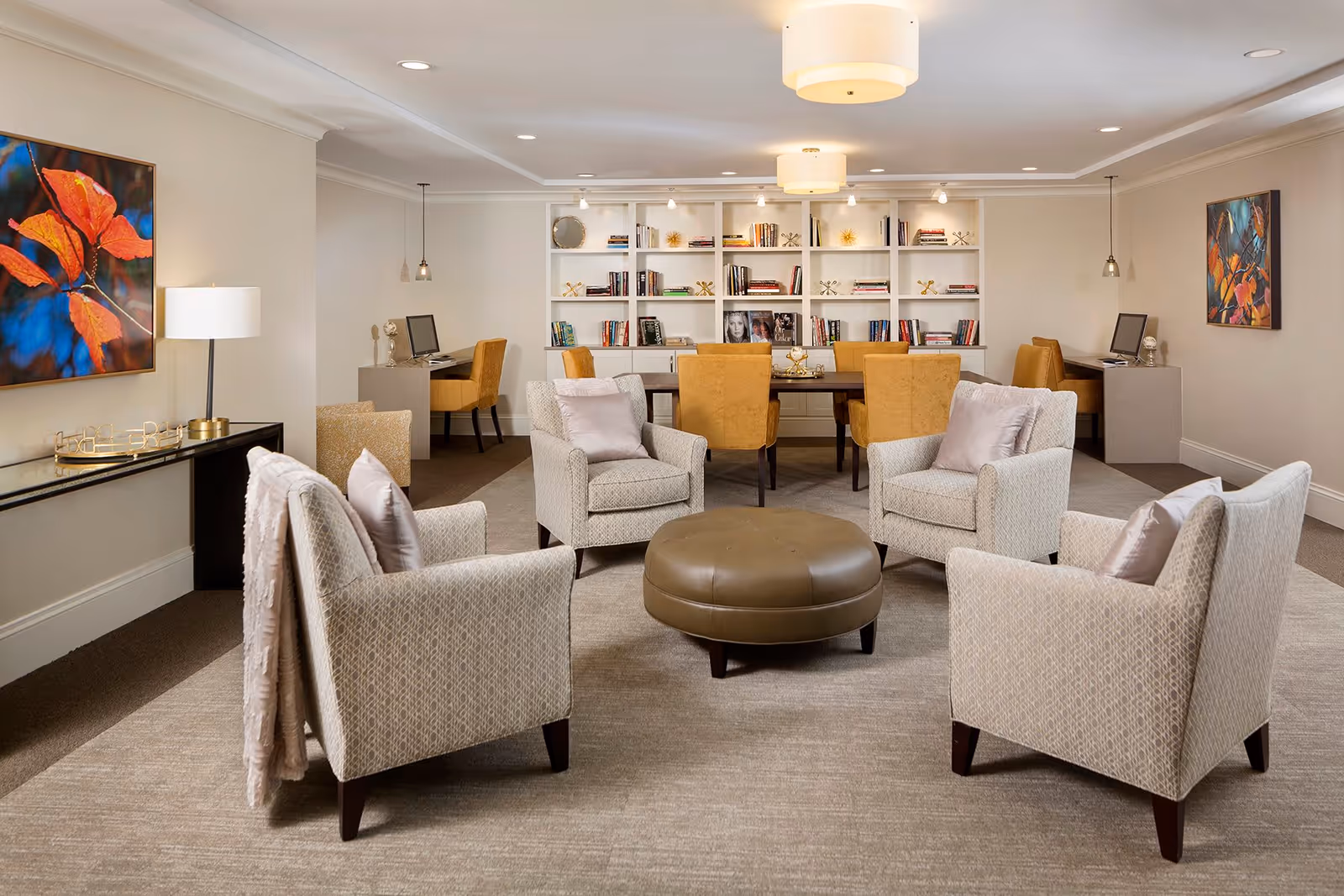 A cozy and well-lit common area with four beige armchairs arranged around a round brown ottoman on a carpeted floor. Behind the seating area is a wooden table with six mustard yellow chairs and a large white bookshelf filled with books and decorative items. Two desks with computers and chairs are positioned against the side walls. The room is decorated with colorful leaf-themed paintings and modern ceiling lights.