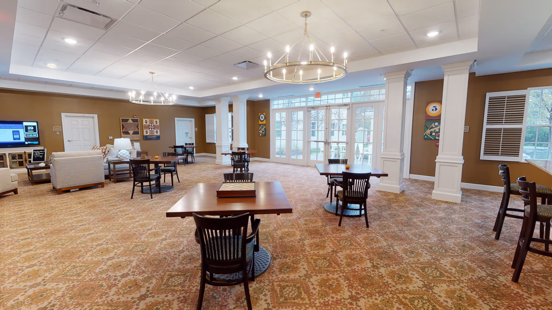 A spacious senior living common area with multiple wooden tables and chairs arranged on a patterned carpet. The room features two large circular chandeliers, beige walls with decorative artwork, and large windows and glass doors letting in natural light. There is a seating area with armchairs and a TV on the left side of the room.