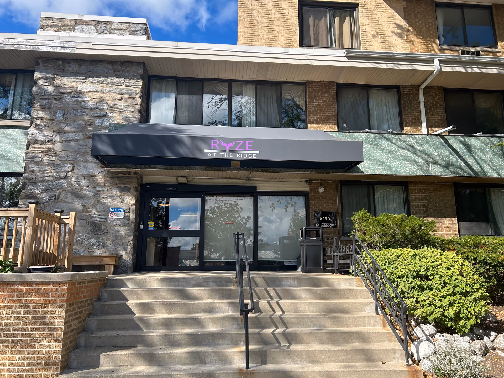 Entrance to a multi-story brick building with stone accents, featuring a black awning with the text 'RYZE AT THE RIDGE' in purple and white. There are concrete steps leading up to glass double doors, with metal railings on either side and greenery on the right side of the steps.