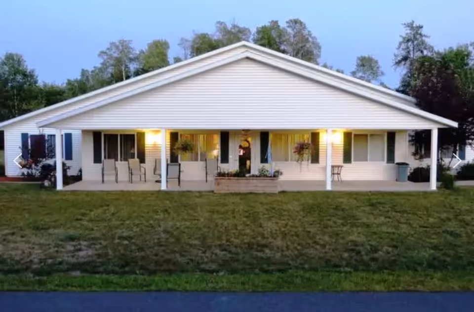 Exterior view of a single-story white building with a covered porch featuring several chairs and hanging flower pots, surrounded by grass and trees in the background during early evening.