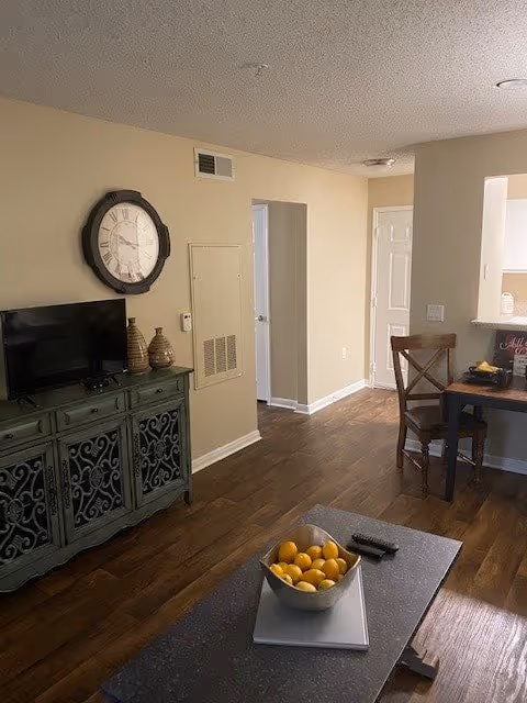 Interior view of a living area in a senior living facility with a wooden floor, a green cabinet holding a TV and decorative vases, a large wall clock above the cabinet, a small dining table with two chairs near a kitchen pass-through window, and a coffee table with a bowl of lemons and a TV remote.