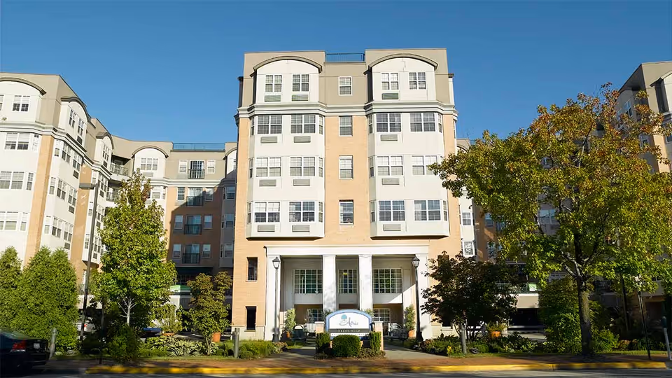 Exterior view of a multi-story senior living facility building with beige and white facade, large windows, and a central entrance with columns. The building is surrounded by trees and landscaping under a clear blue sky.