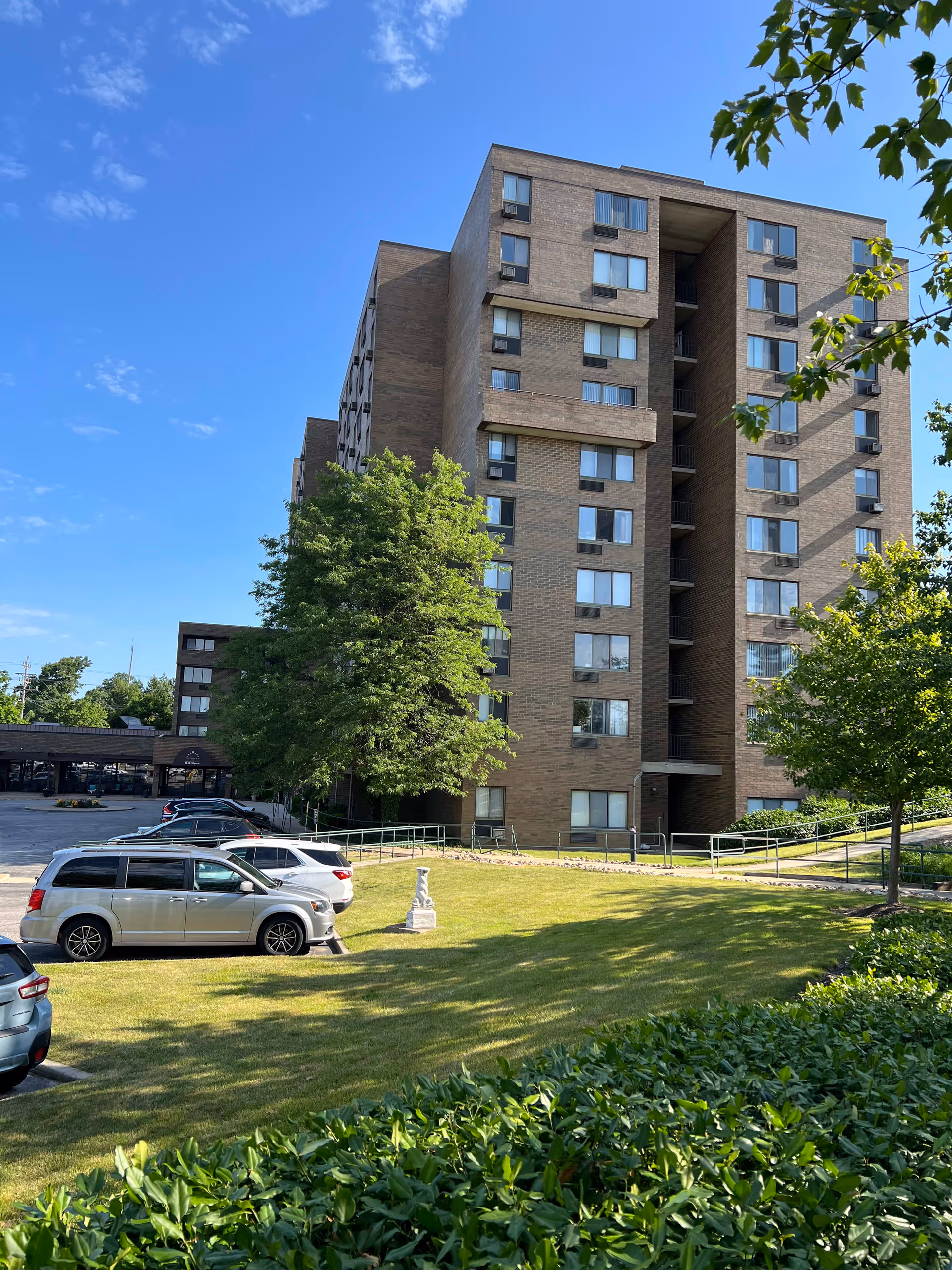 Exterior view of a multi-story brick apartment building with several windows, surrounded by green trees and a grassy lawn. Several cars are parked in a parking lot adjacent to the building under a clear blue sky.