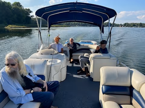 Four elderly people sitting and relaxing on a pontoon boat on a lake with trees and houses visible in the background under a clear blue sky.