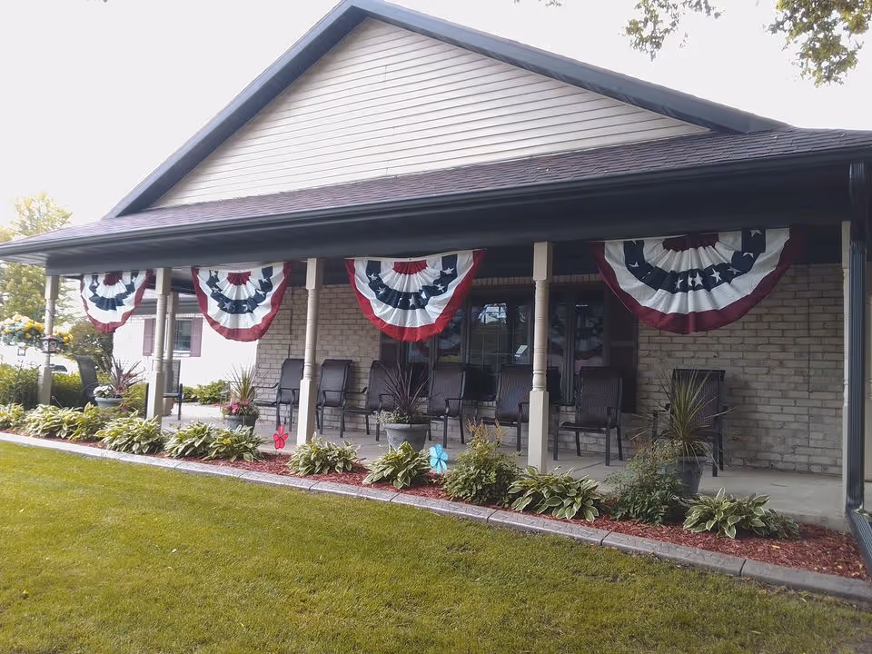 Front porch of a residential building decorated with red, white, and blue patriotic bunting. The porch has several chairs and potted plants, with a well-maintained lawn and garden in front.