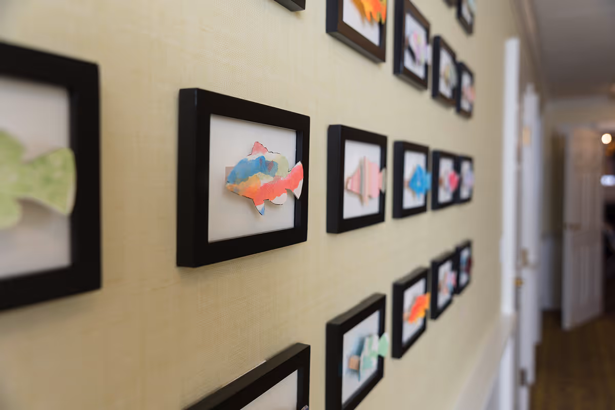 A hallway wall decorated with multiple small black-framed artworks featuring colorful fish cutouts. The hallway has a light-colored wall and a carpeted floor, with doors visible in the background.