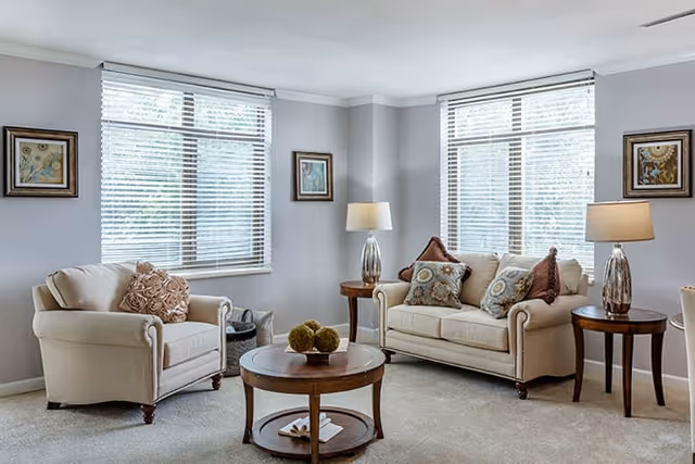 A bright living room with two large windows covered by white blinds. The room features a beige armchair and a matching loveseat, both adorned with decorative pillows. There are two wooden side tables with matching lamps on either side of the loveseat, and a round wooden coffee table with decorative items in the center. The walls are painted light gray and decorated with framed artwork.