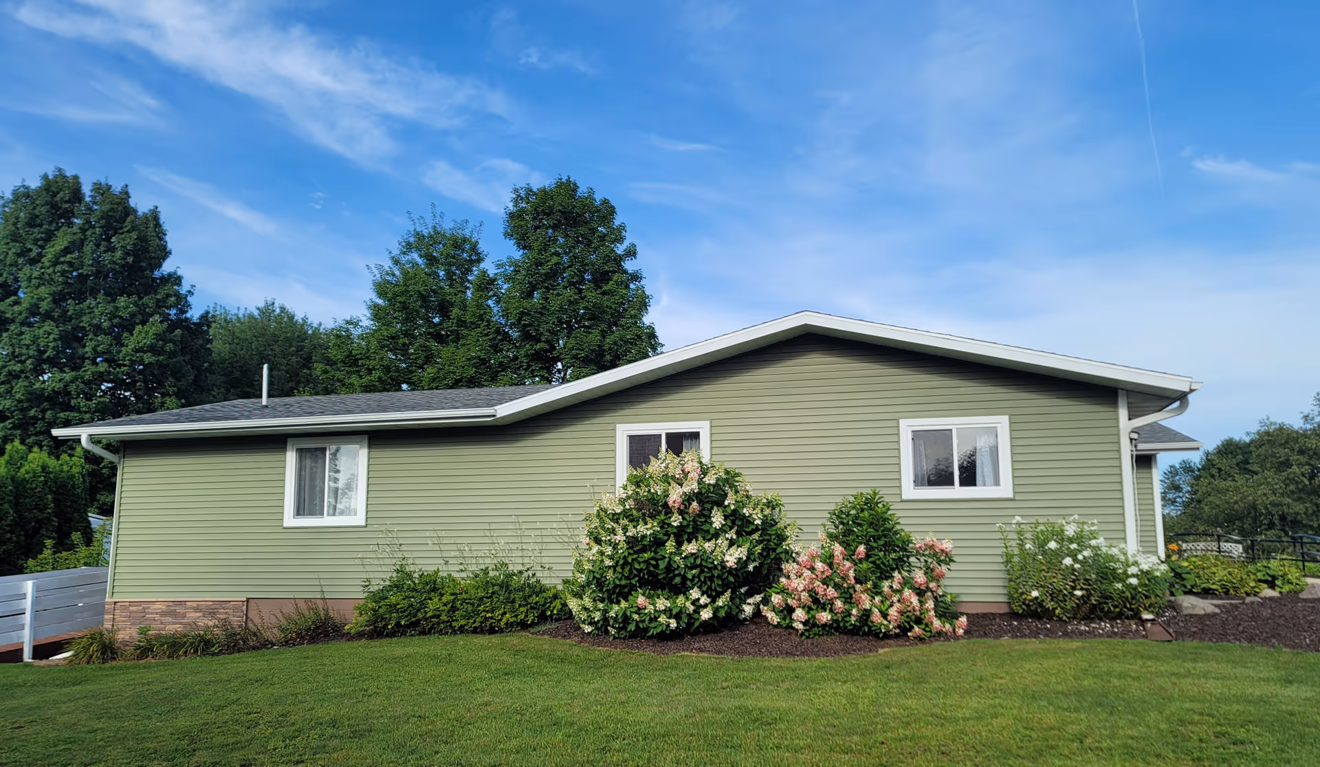 Single-story green building with white trim surrounded by a well-maintained lawn and flowering bushes under a blue sky with some clouds.