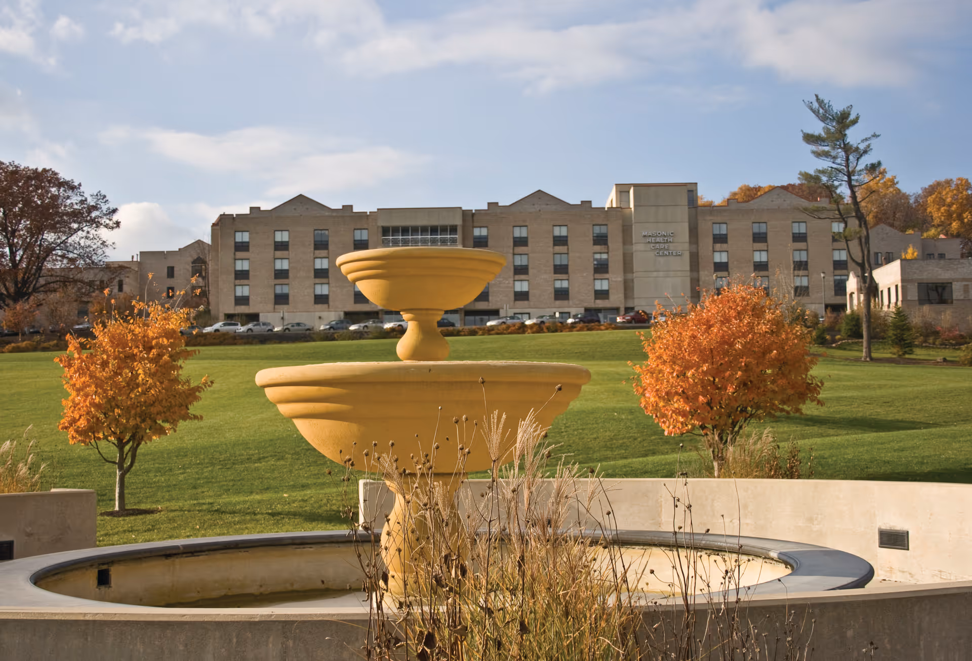 Outdoor view of a senior living facility with a large beige building in the background labeled 'Masonic Health Care Center'. In the foreground, there is a large stone fountain surrounded by a circular concrete structure, with autumn-colored trees and green grass on a sunny day with partly cloudy skies.