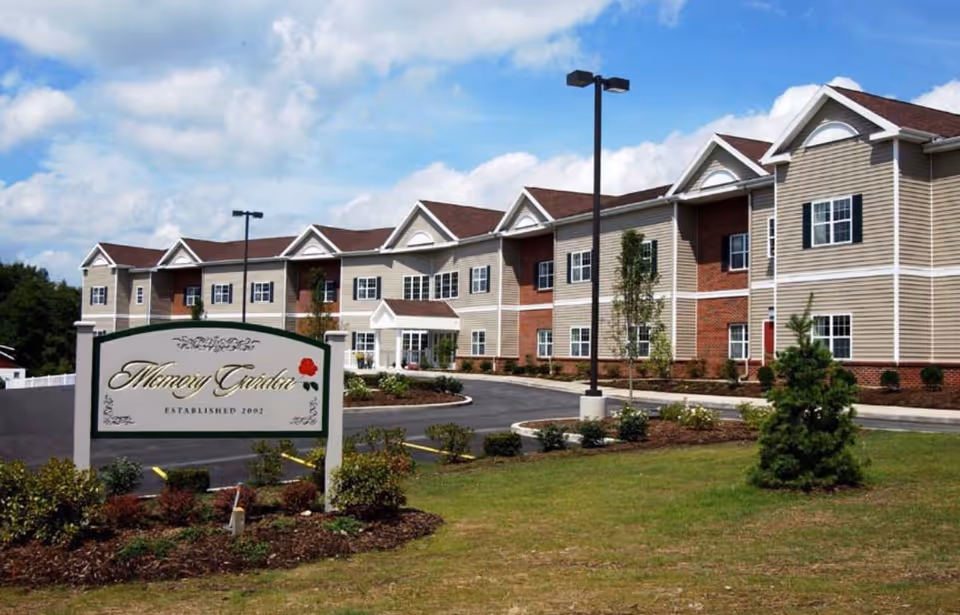 Exterior view of a two-story senior living facility named Memory Garden with a large sign in front, a paved driveway, landscaped bushes, and a partly cloudy sky.