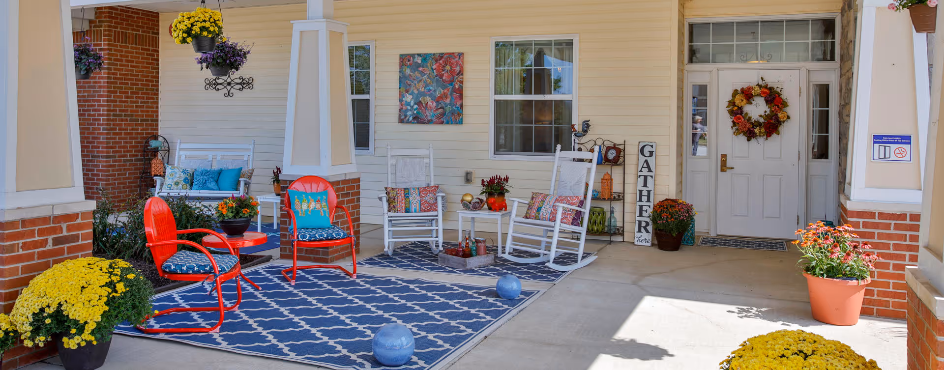 Covered outdoor seating area with red metal chairs, white rocking chairs, a blue patterned rug, potted yellow and orange flowers, and a white door decorated with a fall wreath. The area has brick pillars and a sign that says 'GATHER here'.