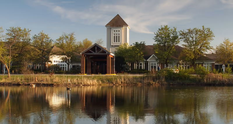 Exterior view of Kendal at Oberlin facility with a pond in the foreground reflecting the building and surrounding trees under a partly cloudy sky.