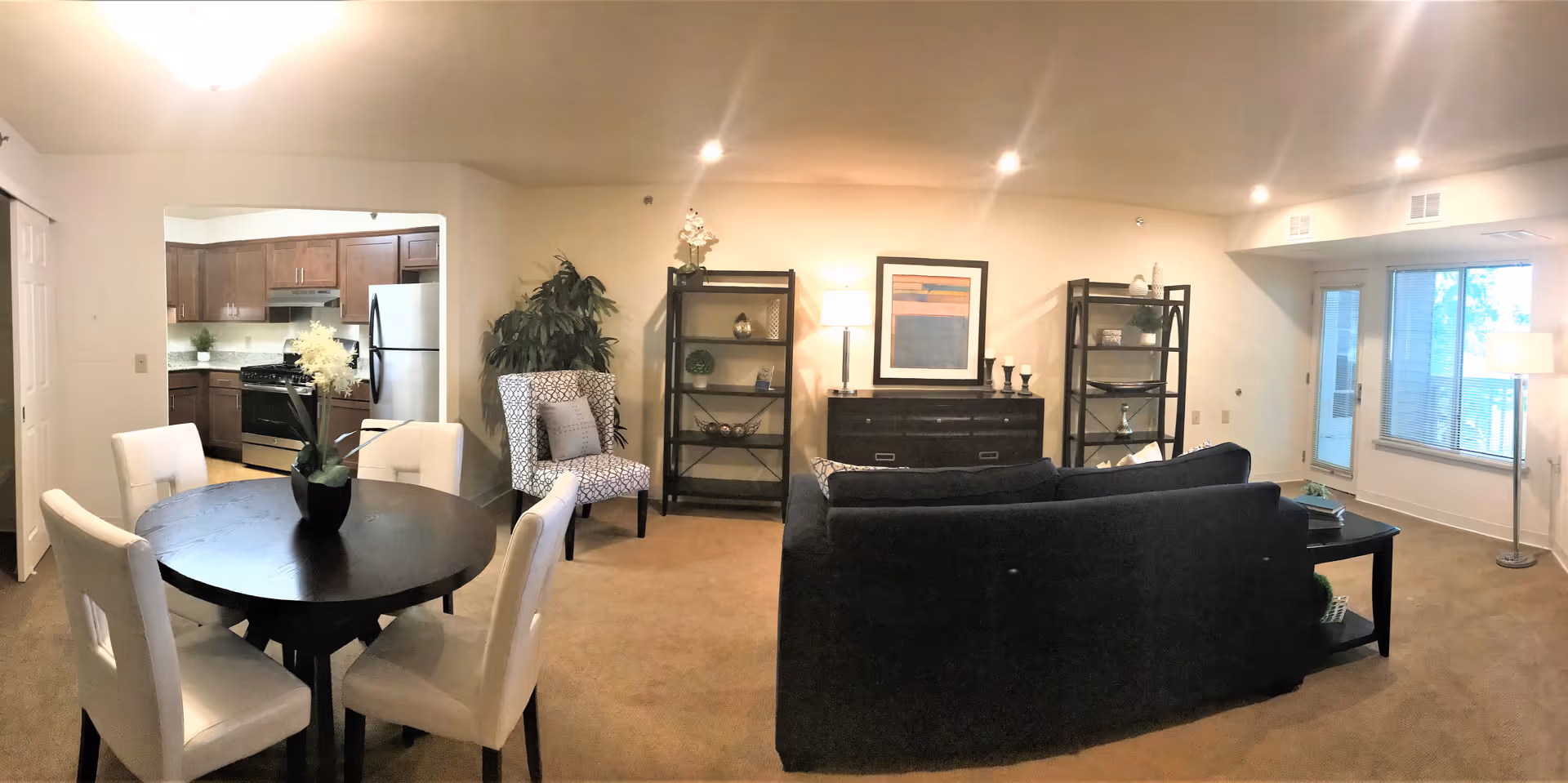 Interior view of a senior living apartment featuring a living room and dining area. The living room has a dark sofa, two black shelving units, a sideboard with a lamp and framed artwork, and a patterned armchair next to a large plant. The dining area has a round dark wooden table with four white upholstered chairs and a flower vase centerpiece. The kitchen with wooden cabinets and stainless steel appliances is visible through an open doorway. Large windows and a glass door provide natural light to the space.