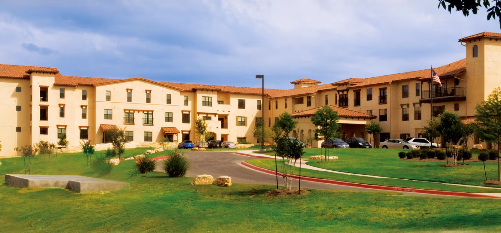 Exterior view of Buckner Villas, a large multi-story senior living facility with beige walls and red-tiled roofs, surrounded by green lawns, small trees, and a curved driveway with parked cars.