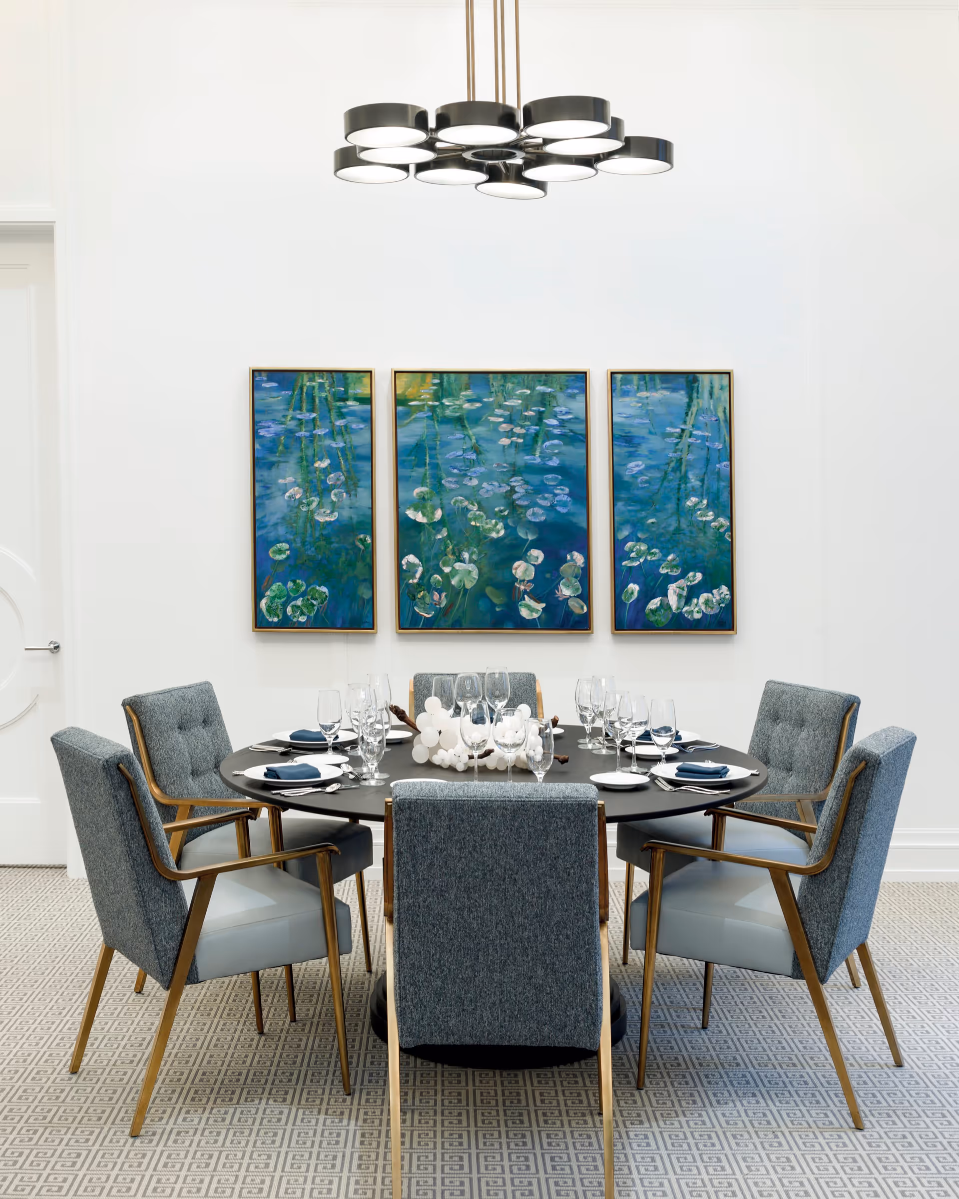 A modern dining room with a round black table set for six people. The table is surrounded by six upholstered chairs with wooden legs and armrests. On the table are neatly arranged plates, napkins, and glassware. Above the table hangs a contemporary black chandelier with multiple circular lights. On the white wall behind the table are three framed paintings depicting water lilies on a blue water surface. The floor has a patterned carpet in neutral tones.