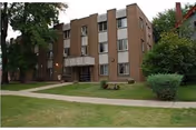 A three-story brick building with multiple windows, surrounded by green grass, trees, and bushes, with a concrete walkway leading to the entrance.