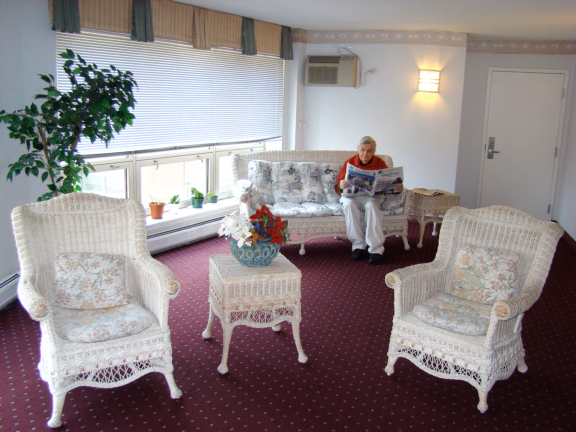 A senior man sitting on a white wicker sofa reading a newspaper in a bright room with large windows covered by blinds. The room has two matching white wicker armchairs with floral cushions, a wicker side table with a flower arrangement, a potted plant near the window, and a maroon carpet with a small pattern.
