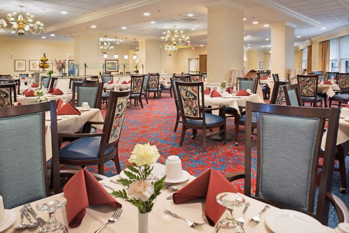 A spacious dining room with multiple tables set with white tablecloths, red folded napkins, white cups, plates, and silverware. The chairs have dark wooden frames with patterned upholstery on the backrests. The room features a red and blue patterned carpet, chandeliers, and soft yellow walls with decorative columns. There are floral centerpieces on the tables and large windows with curtains along one side of the room.