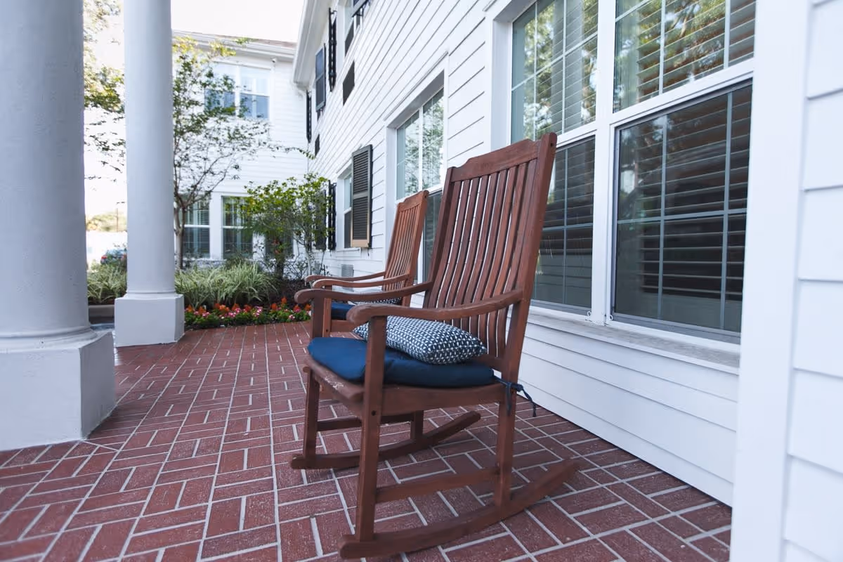 Two wooden rocking chairs with blue cushions and patterned pillows on a red brick patio outside a white building with large windows and black shutters. There are plants and flowers along the edge of the patio.