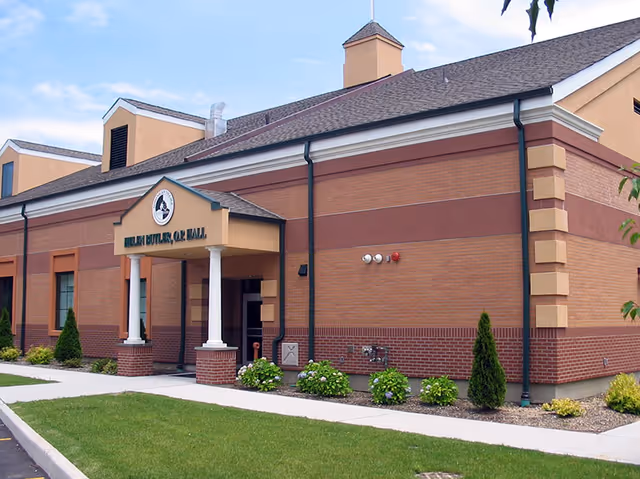 Exterior view of a brick building with a small covered entrance supported by two white columns. The building has a sign above the entrance that reads 'HELEN BUTLER, O.A.R. HALL'. There are small bushes and plants along the side of the building and a well-maintained lawn in front.