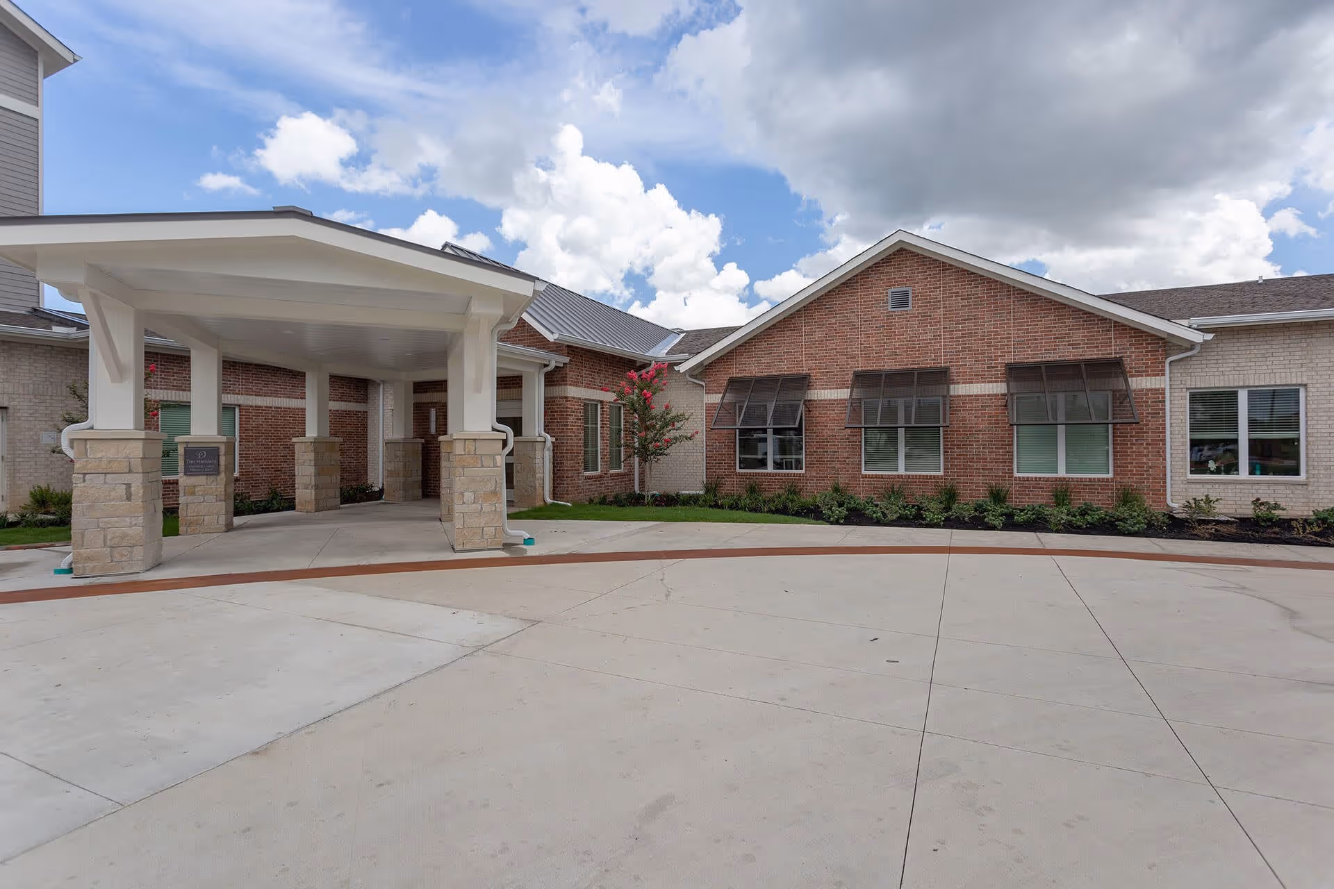 Front exterior view of a senior living facility building with a covered entrance supported by stone pillars, brick and light-colored siding walls, several windows with awnings, and a concrete driveway under a partly cloudy sky.