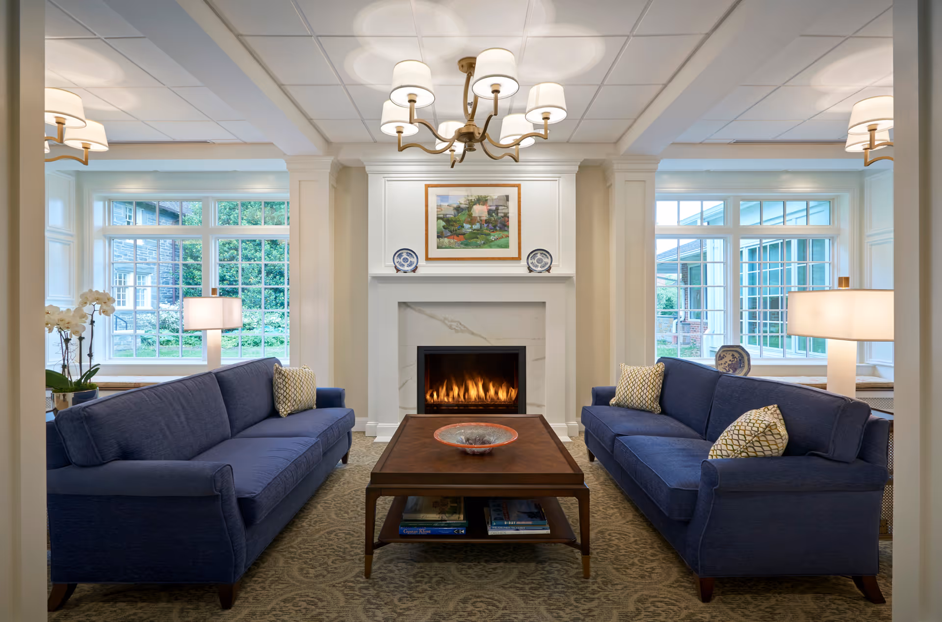 A cozy living room with two blue sofas facing each other across a wooden coffee table. The room features a lit fireplace with a framed painting and decorative plates above it. Large windows on either side let in natural light, and there are table lamps and a chandelier providing additional lighting. The room has a warm and inviting atmosphere with neutral-colored walls and carpet.