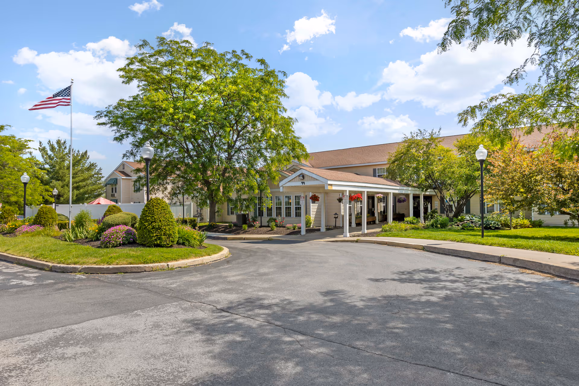 Exterior view of Brookdale Clinton IL senior living facility with a driveway, landscaped garden with bushes and flowers, an American flag on a flagpole, and a covered entrance to the building under a partly cloudy sky.