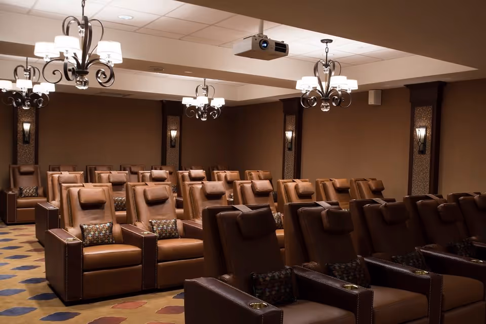 A cozy theater room with multiple rows of brown leather recliner chairs, each with a small decorative pillow. The room has warm lighting from chandeliers and wall sconces, and a ceiling-mounted projector is visible.