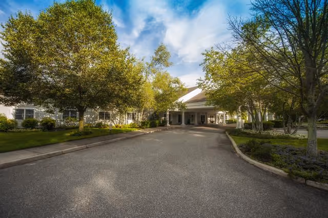 Driveway leading to the entrance of a building surrounded by trees and greenery under a partly cloudy sky.
