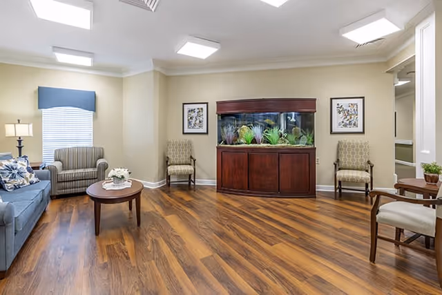 A senior living facility common area with wooden flooring, a blue sofa with patterned cushions, a round wooden coffee table with a flower arrangement, two patterned armchairs, and a large fish tank set in a wooden cabinet against a beige wall. Two framed pictures hang on the wall beside the fish tank, and a window with a blue valance is visible on the left side.