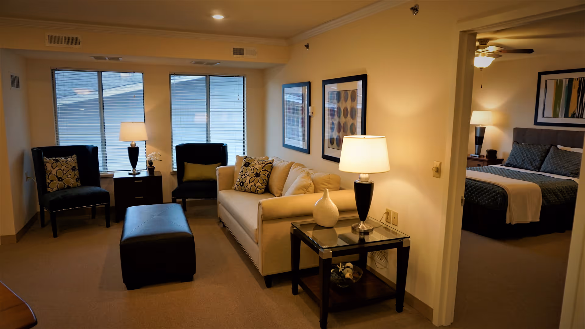 A cozy living room area with a beige sofa, two dark armchairs with patterned cushions, a black ottoman, and two side tables with lamps. The room has three large windows with blinds. Through an open doorway, a bedroom with a bed, nightstand, lamp, and framed artwork is visible.