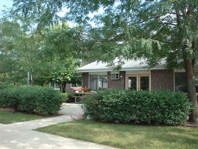 A single-story brick building partially obscured by green bushes and trees, with a concrete walkway leading to the entrance. The scene is outdoors with a well-maintained lawn and shaded by tree branches.