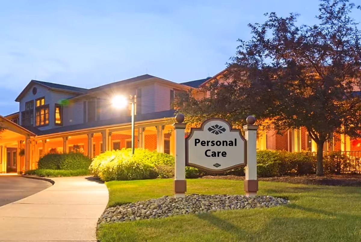 Exterior view of a senior living facility building at dusk with warm lights glowing from inside. A sign in the foreground reads 'Personal Care' and is surrounded by landscaping with bushes and a tree.