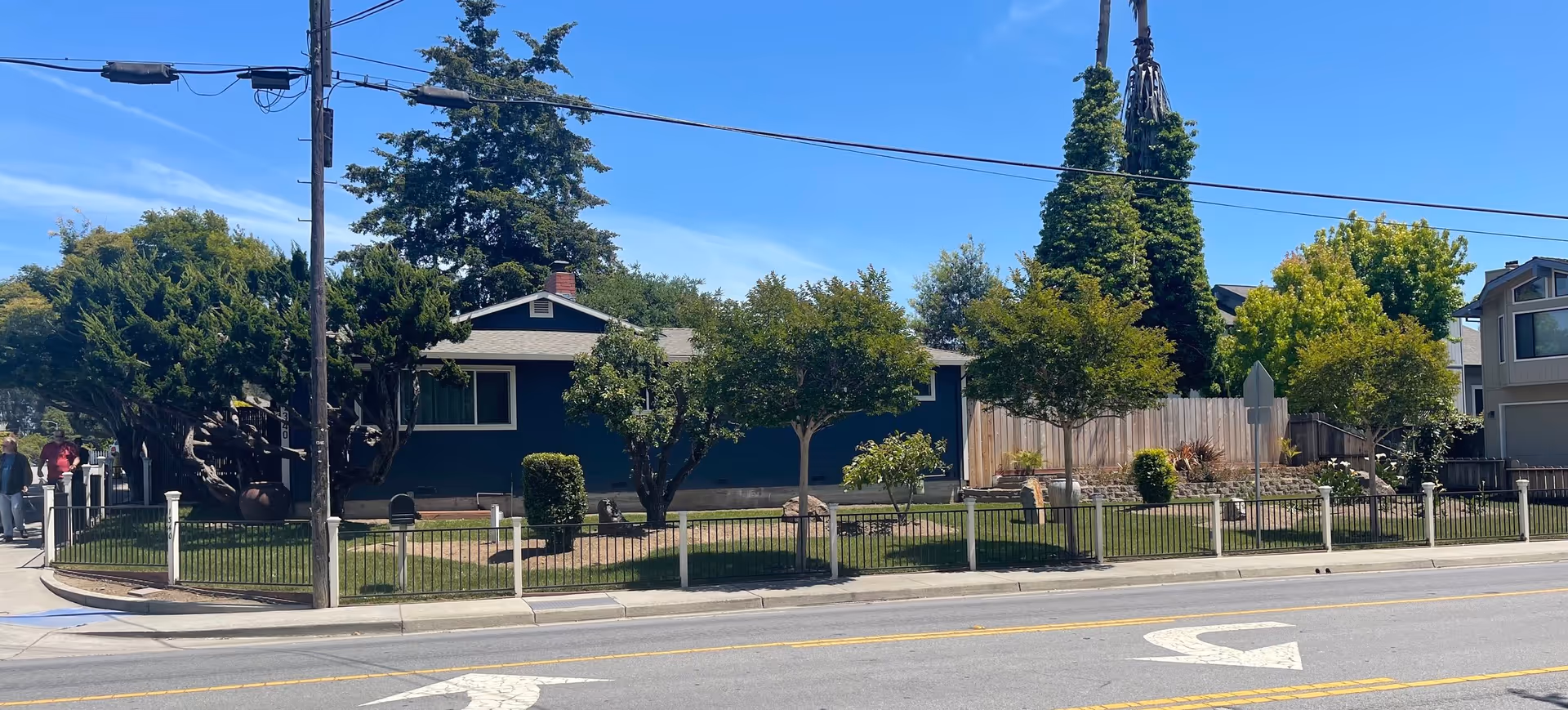 Exterior view of a single-story residential building with a dark blue facade, surrounded by a black metal fence and several trees and shrubs in the front yard. The building is located along a street with visible road markings and utility poles. The sky is clear and blue.