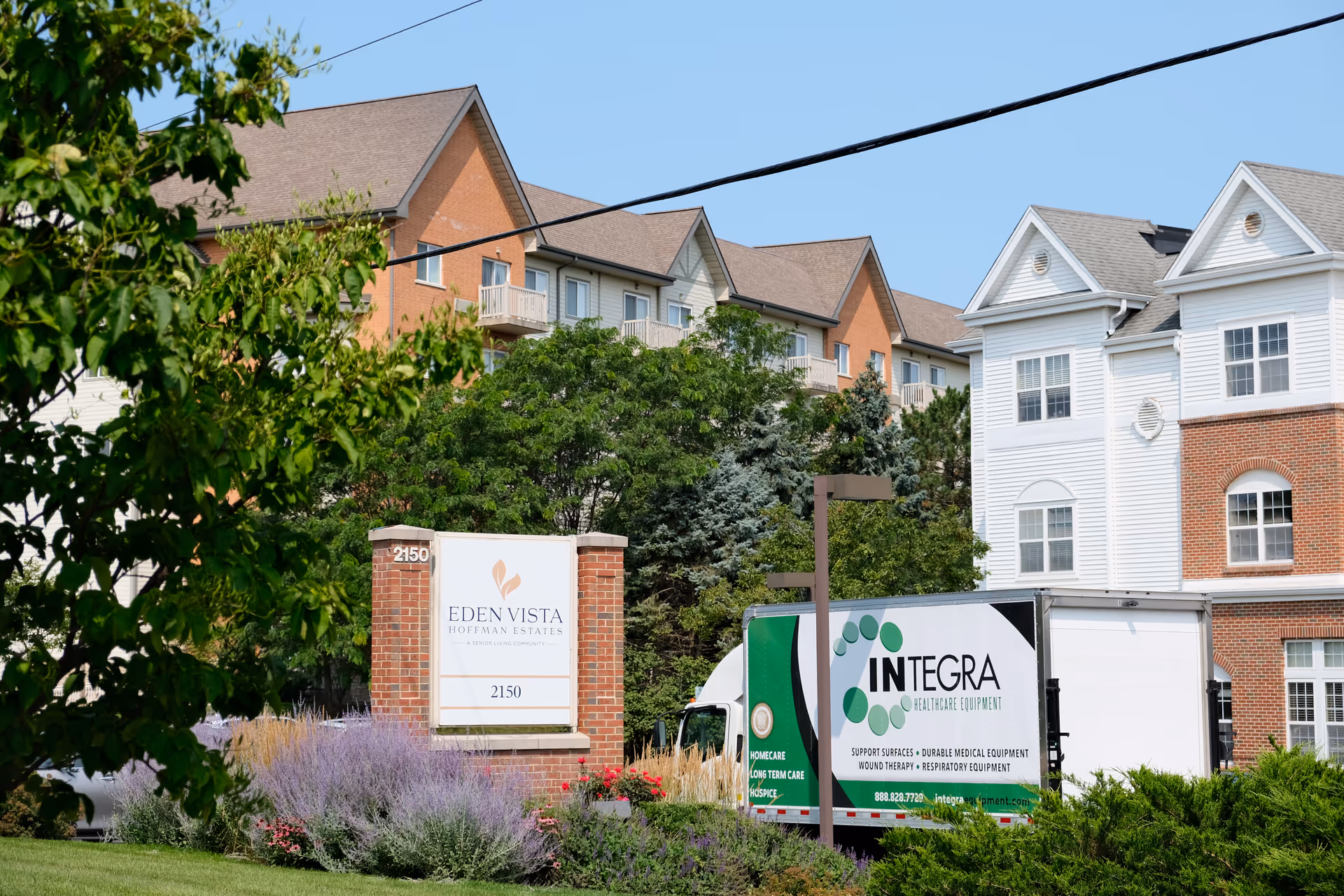 Exterior view of Eden Vista Hoffman Estates senior living community with a brick and white building, green trees, and a sign displaying the facility name and address 2150. A delivery truck from Integra Healthcare Equipment is parked nearby.