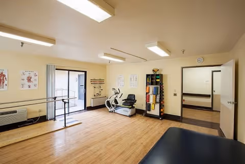 A physical therapy or exercise room with wooden flooring, parallel bars for walking support, an exercise bike, a rack holding various exercise balls and foam rollers, anatomical charts on the walls, and a treatment table in the foreground. The room has fluorescent ceiling lights and a door leading to another area.