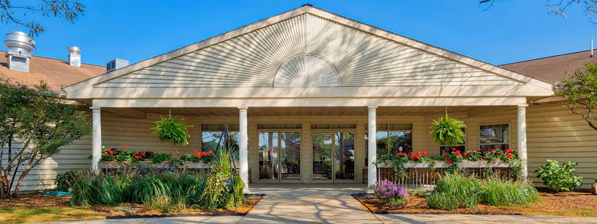 Front exterior view of a single-story building with a peaked roof, beige siding, and a covered entrance supported by white columns. There are hanging green plants and flower boxes with red and purple flowers along the front porch. Trees and shrubs surround the building under a clear blue sky.