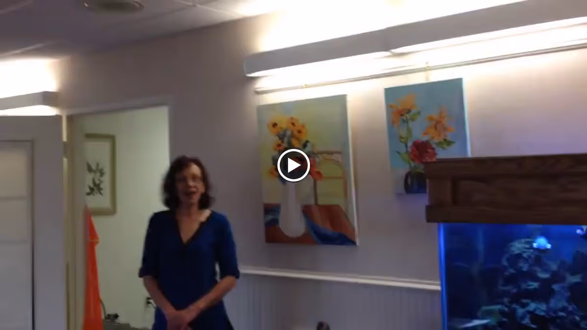 An indoor room with a woman standing near a doorway. The wall behind her has two colorful paintings of flowers, and there is a large aquarium with blue lighting on the right side of the image.
