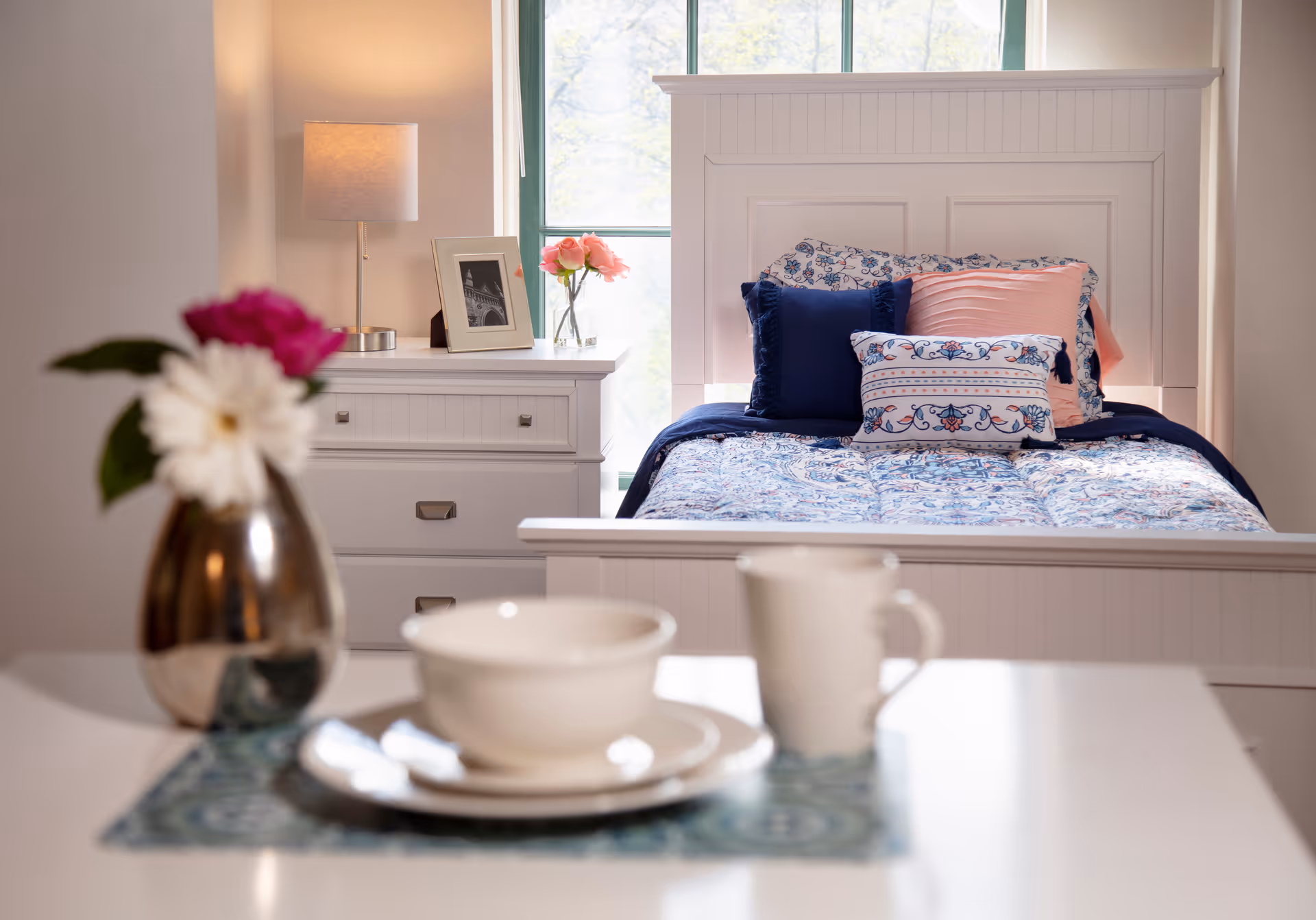 A cozy bedroom with a white wooden bed featuring a patterned blue and white bedspread and several decorative pillows in blue and pink. Next to the bed is a white dresser with a lamp, a framed photo, and a vase with pink flowers. In the foreground, there is a table set with a white bowl, plate, and mug, along with a small vase holding a pink and white flower.