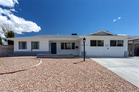 Single-story white building with a gray roof under a blue sky with some clouds. The front yard is covered with gravel and has a concrete driveway on the right side. There is a black lamp post near the center of the yard and several windows on the building's front facade.
