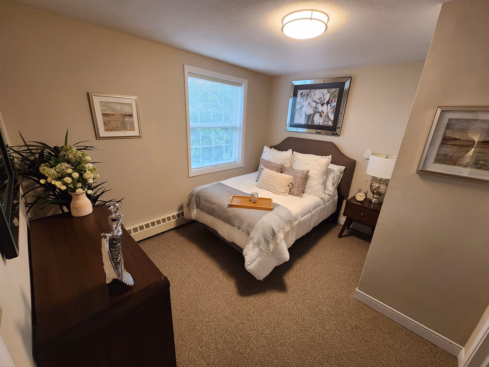 A cozy bedroom with a neatly made bed featuring white and gray bedding and multiple pillows. A wooden tray with a cup is placed on the bed. The room has beige walls, a window with blinds, a ceiling light, a wooden dresser with decorative items including a vase with flowers, and a nightstand with a lamp and an alarm clock. Two framed pictures hang on the walls.