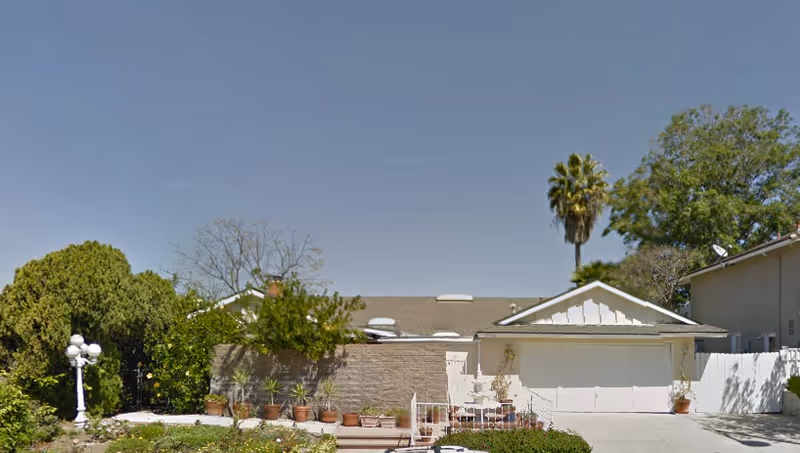 Front exterior view of a single-story residential building with a garage, surrounded by trees and plants under a clear blue sky.