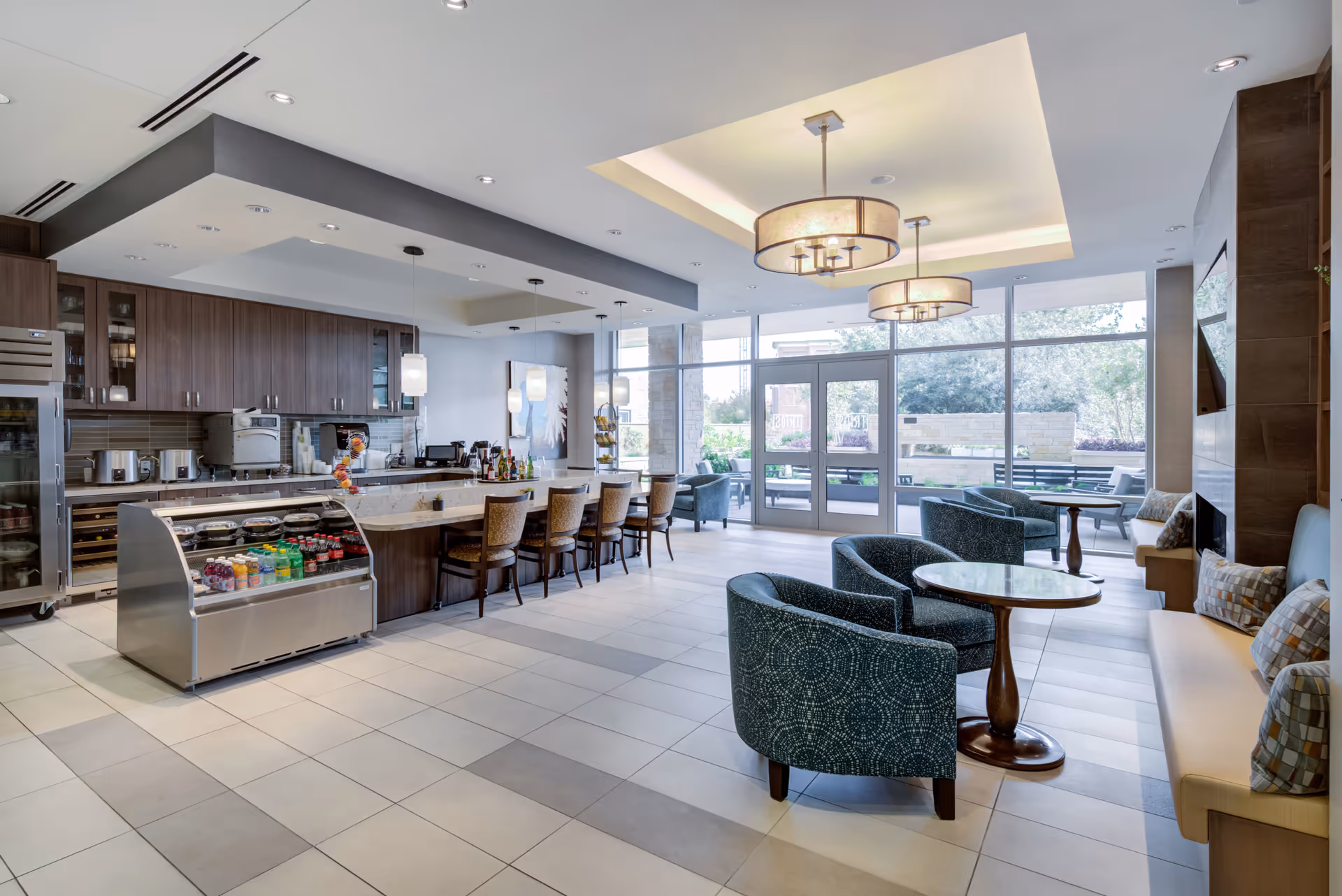 A modern communal lounge area with a kitchen counter and bar stools on the left, a refrigerated display case with beverages, and seating arrangements including blue patterned armchairs and cushioned benches with pillows. Large windows and glass doors provide natural light and a view of an outdoor patio area.